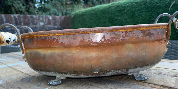rustic copper-colored metal planter or decorative bowl with a weathered patina, featuring small feet at the bottom and looped handles on both sides, shown on a wooden deck with a garden background.