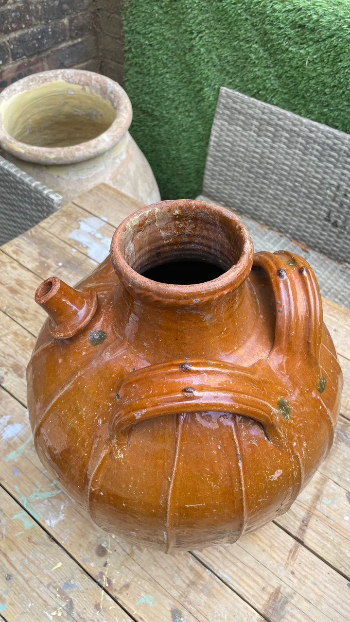 A large rustic glazed ceramic jug with a rounded body, side spout, and handle, shown on a wooden table with patio furniture in the background.