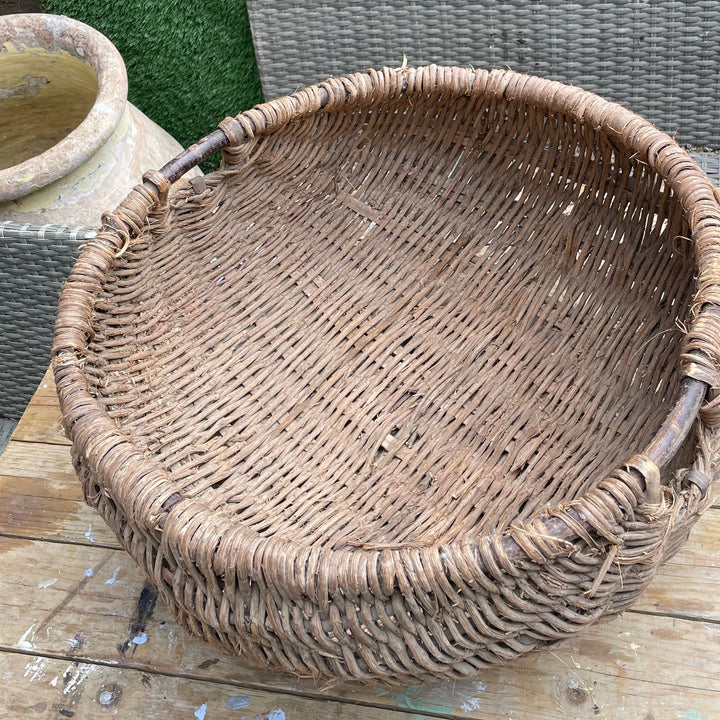 A large round woven wicker basket with dark brown handles, resting on a weathered wooden deck in front of a green hedge.
