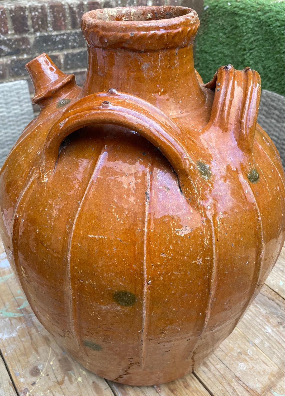 A large glazed ceramic jug with a rounded body, narrow neck, and side spouts, shown on a wooden surface outdoors.