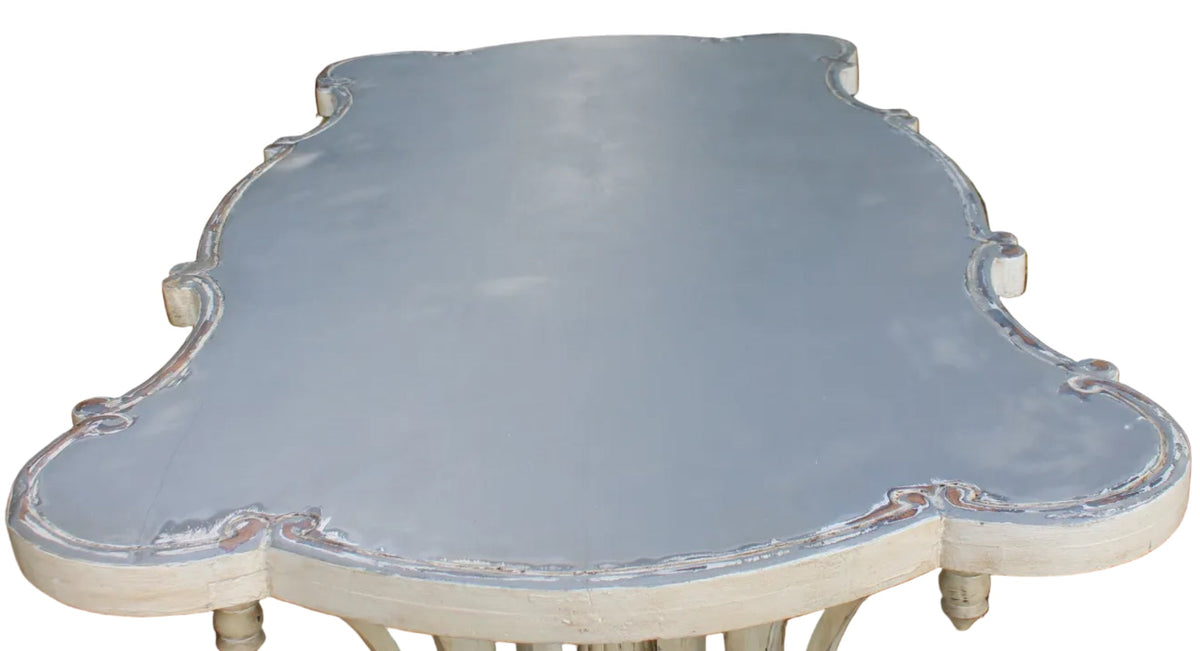 A distressed outdoor garden table with a light gray painted top and an off-white carved pedestal base