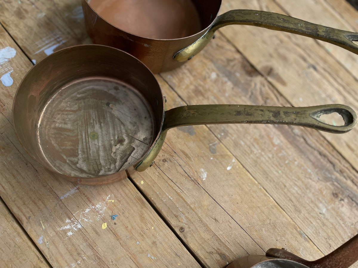 A close-up of a small copper saucepan with a tarnished brass-colored handle resting on a rustic wooden table, with another copper pot partially visible in the background.