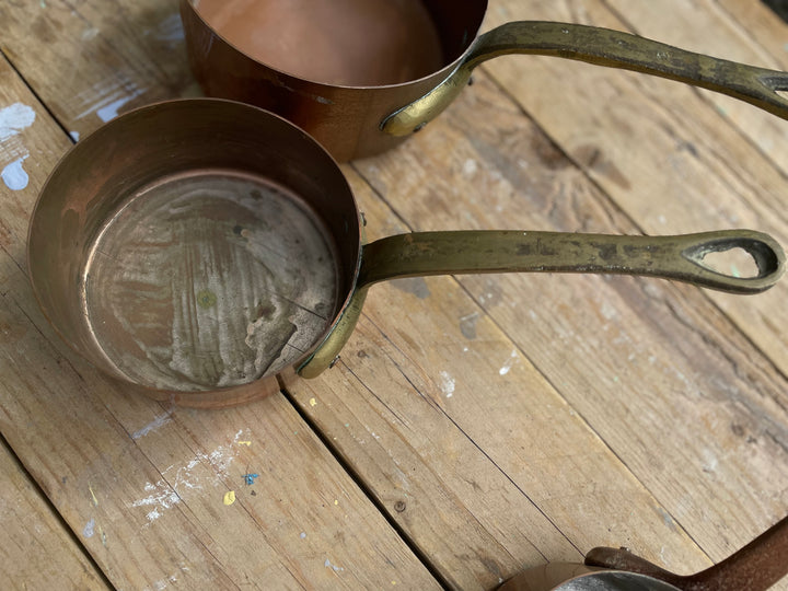 A close-up of a small copper saucepan with a tarnished brass-colored handle resting on a rustic wooden table, with another copper pot partially visible in the background.