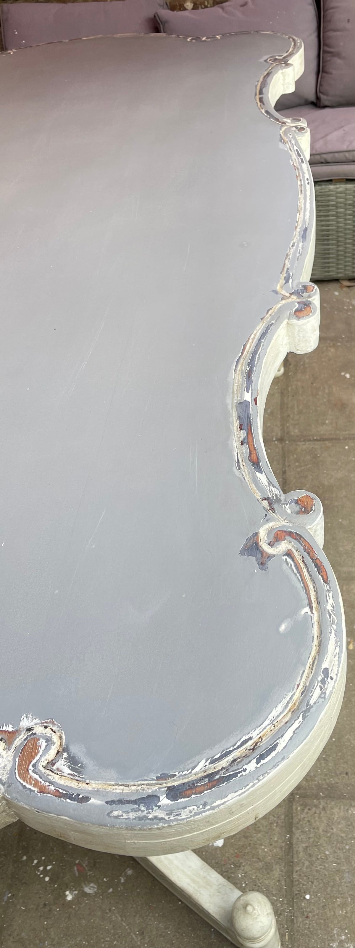A close-up of a distressed painted wooden table corner with a light gray finish and chipped white trim, set on a tiled floor next to a wicker sofa.
Details