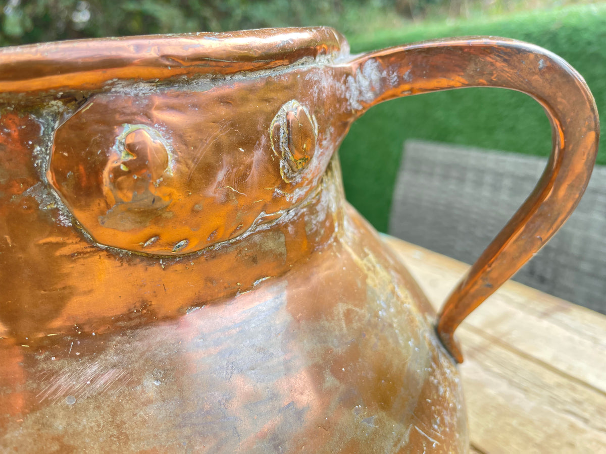 A close-up of a hammered copper pitcher with a curved handle, resting on a wooden table with a blurred green garden background.