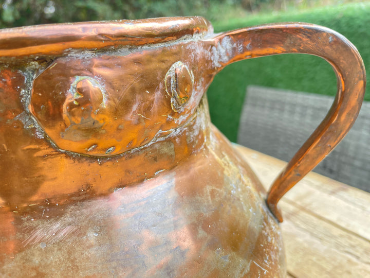A close-up of a hammered copper pitcher with a curved handle, resting on a wooden table with a blurred green garden background.