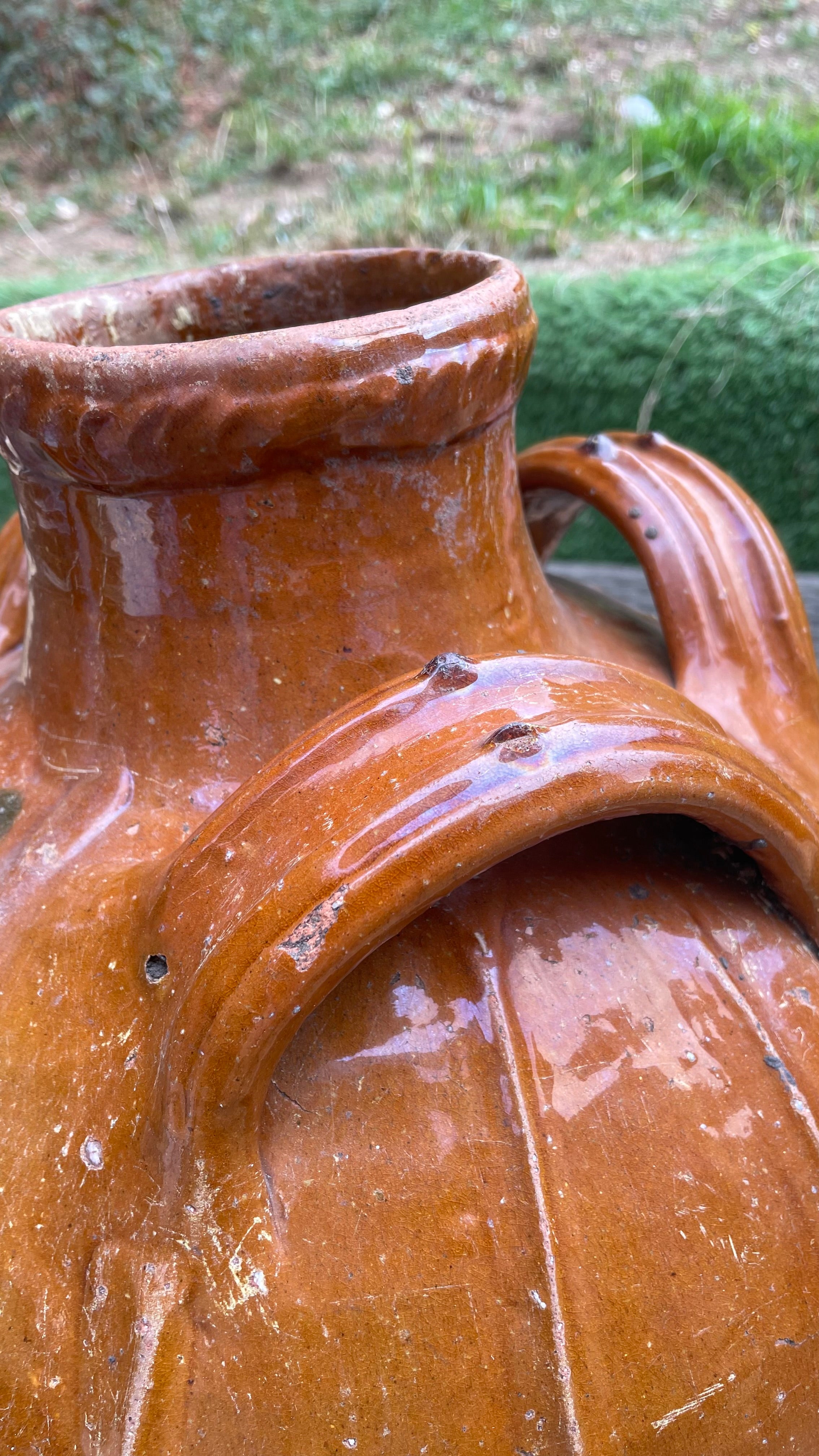 Close-up of a large rustic ceramic vase or jug with a glossy terracotta brown glaze, showing a wide flared rim and a curved handle, photographed outdoors with blurred greenery in the background.