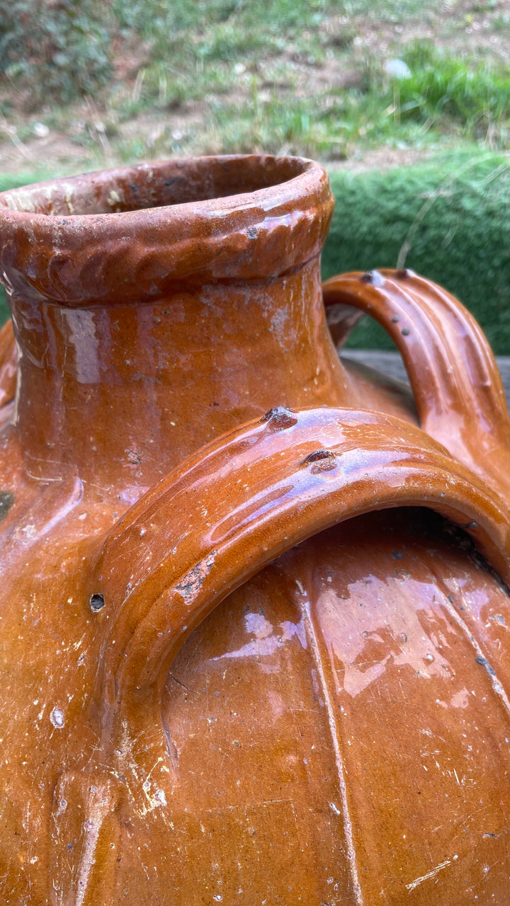 Close-up of a large rustic ceramic vase or jug with a glossy terracotta brown glaze, showing a wide flared rim and a curved handle, photographed outdoors with blurred greenery in the background.