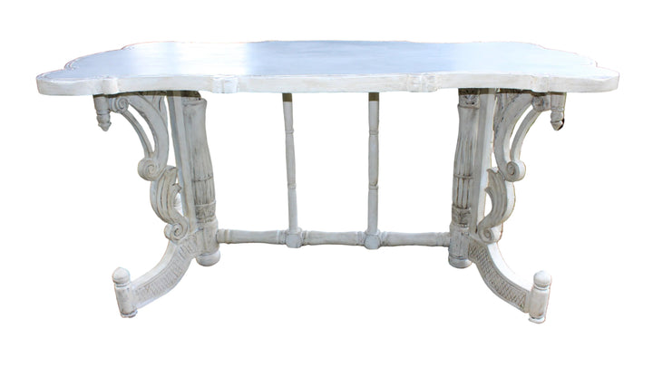 A distressed white outdoor dining table with ornate turned legs and a scalloped apron