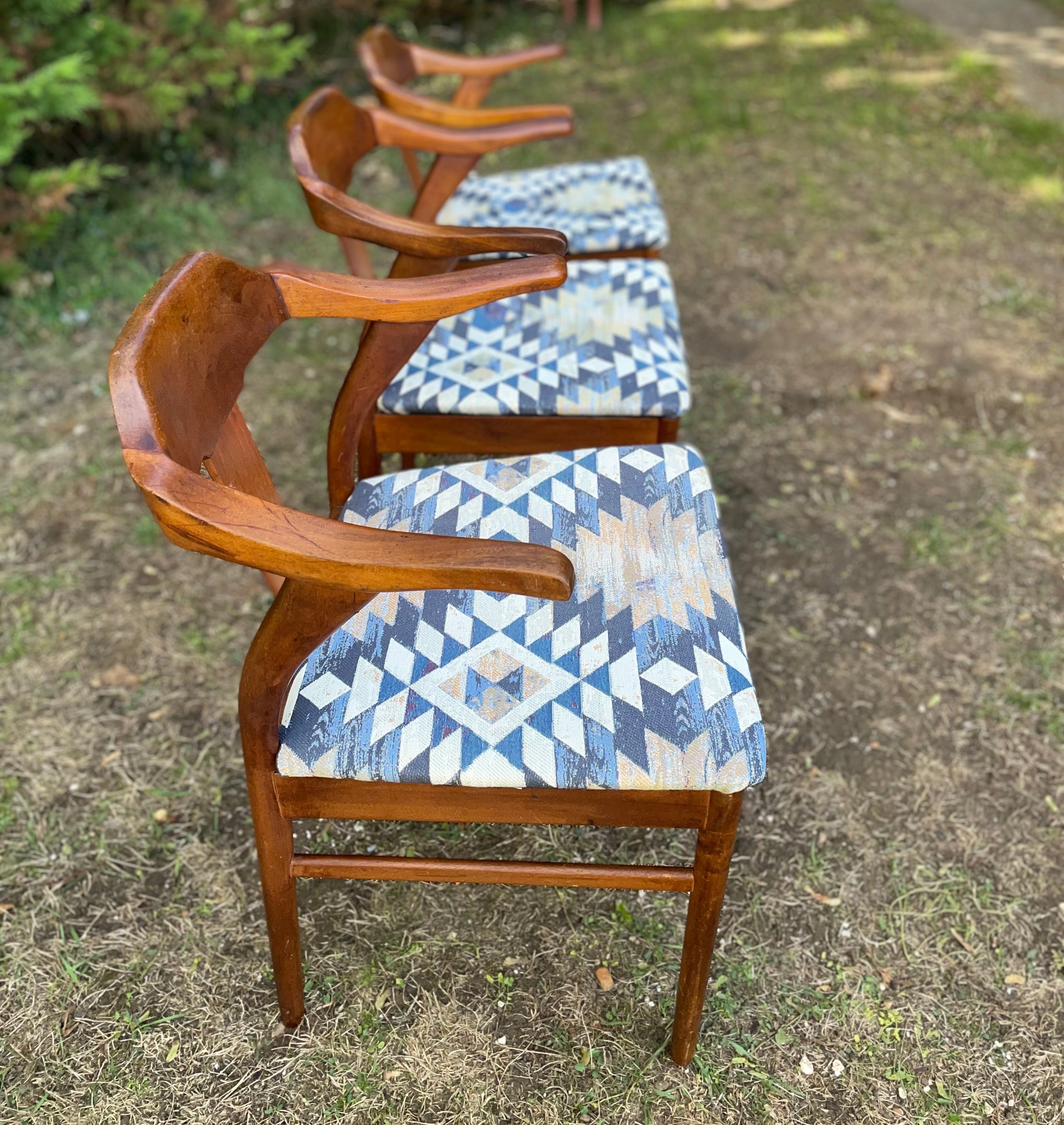 Four mid-century modern wooden armchairs with blue and beige geometric patterned upholstery, arranged in a row on a grassy outdoor lawn.
