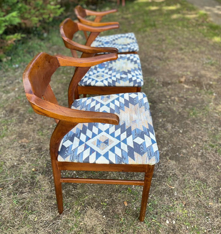 Four mid-century modern wooden armchairs with blue and beige geometric patterned upholstery, arranged in a row on a grassy outdoor lawn.