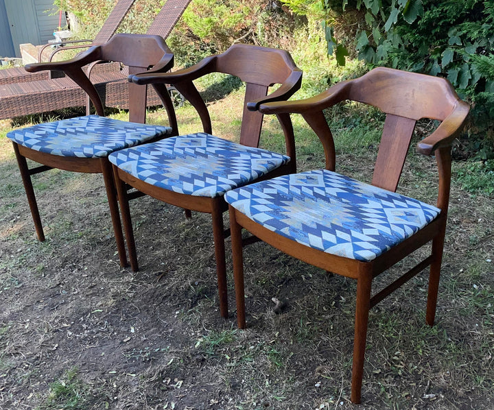 Three mid-century modern wooden dining chairs with curved armrests and blue patterned seat cushions, arranged on a grassy lawn outdoors.