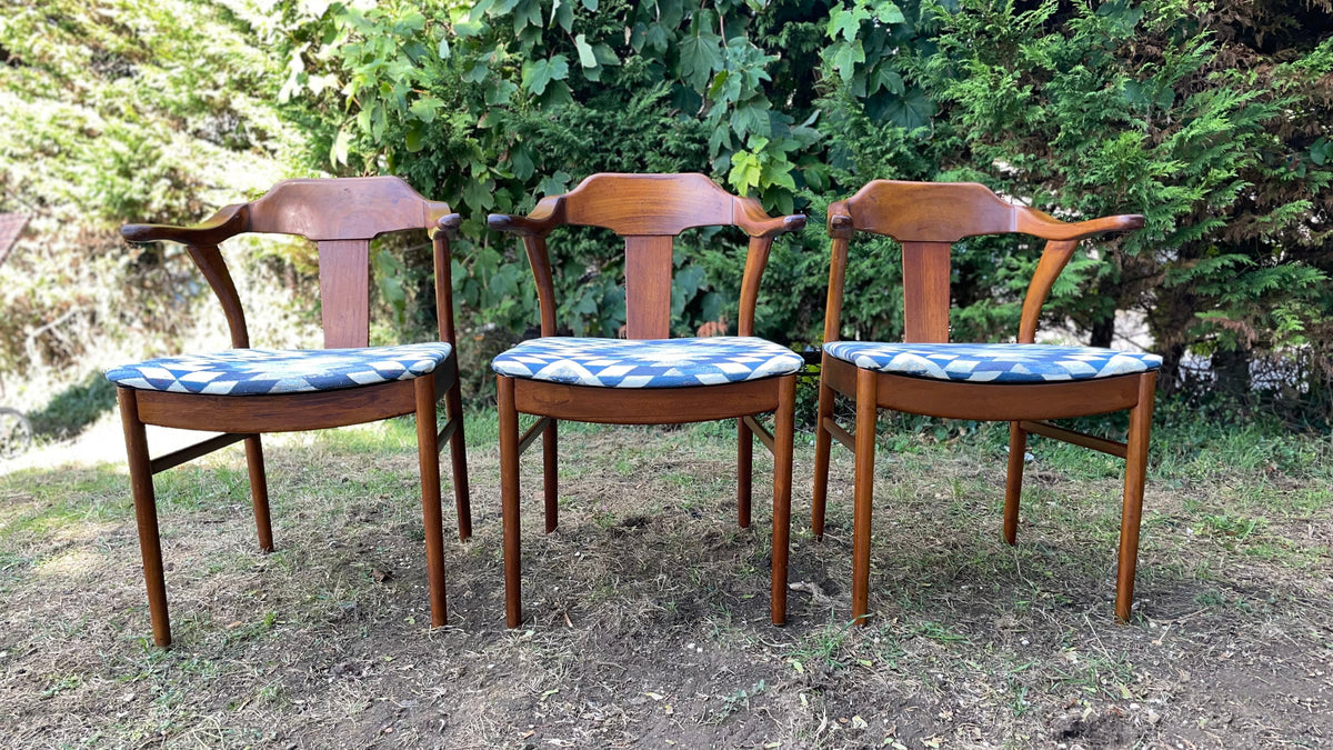 Three mid-century modern wooden armchairs with blue patterned seat cushions, set outdoors on a patch of grass and dirt with a green hedge background.