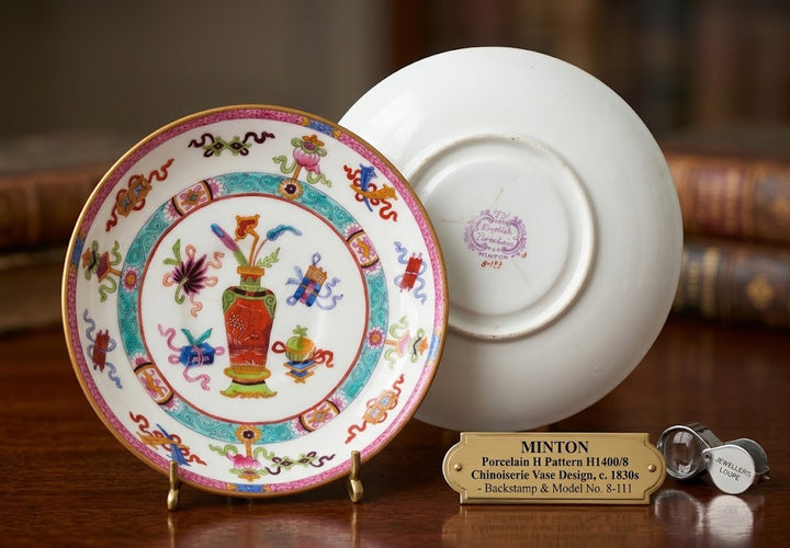 A Minton porcelain pattern H1400/R chinaware vase design saucer, displayed on a wooden table with a second saucer showing the back and a small gold-colored metal plaque.