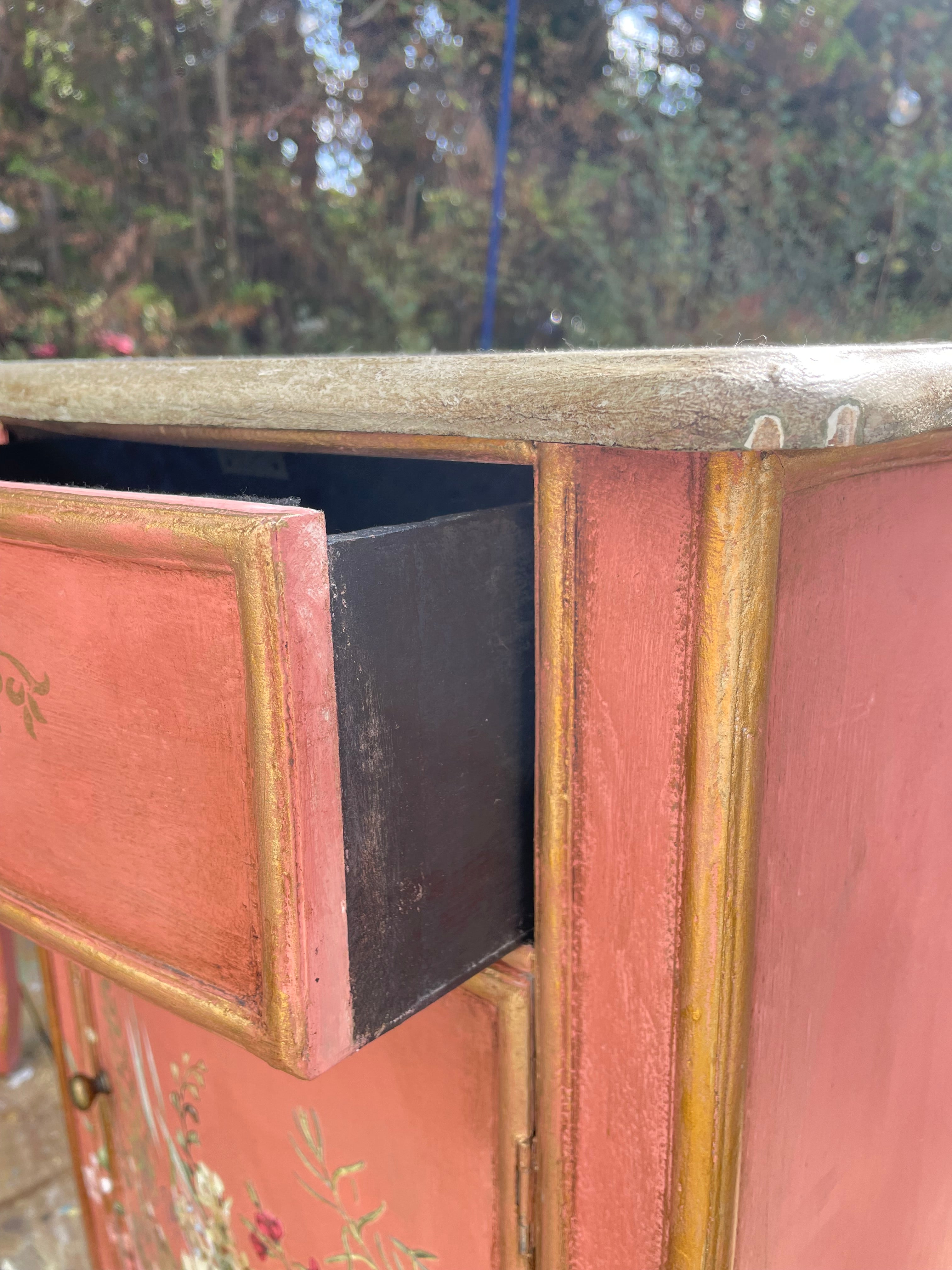 Close-up of a distressed red and gold painted wooden cabinet with one drawer pulled open, showing a dark interior, set against a blurred outdoor background of trees.