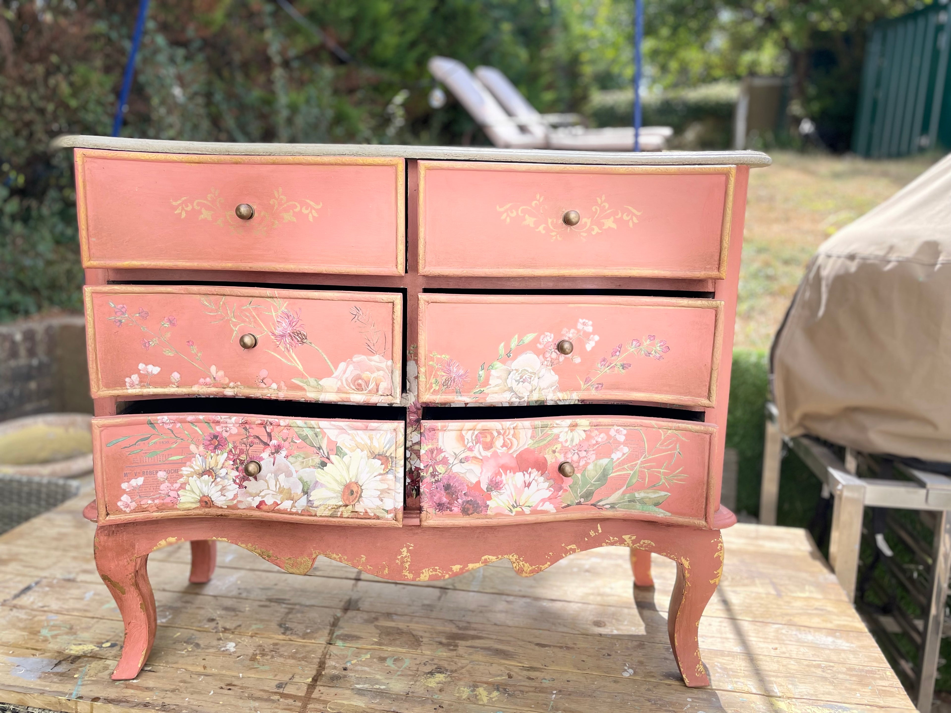 A six-drawer painted wooden chest of drawers with floral decals, set on a wooden deck in an outdoor backyard.