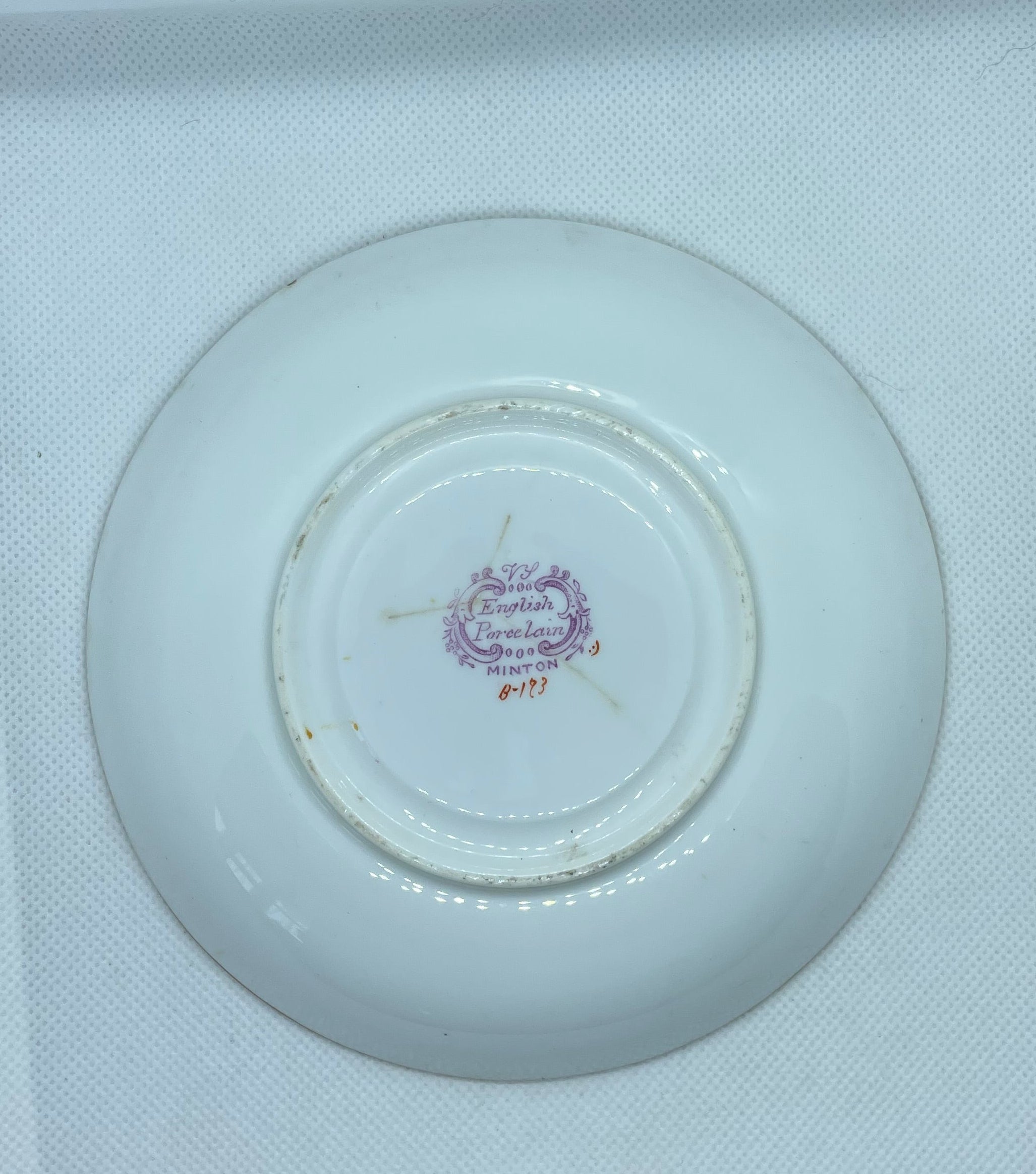 white porcelain saucer or small plate shown from the bottom side, resting inside a white textured box. The center features a small purple and gold printed emblem.
Details