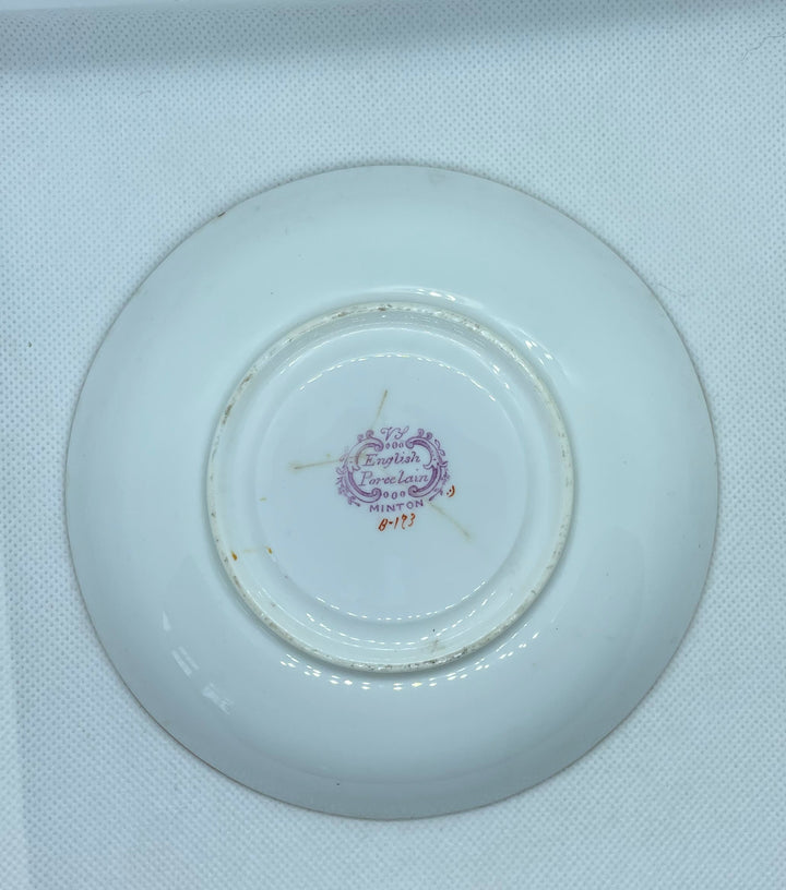 white porcelain saucer or small plate shown from the bottom side, resting inside a white textured box. The center features a small purple and gold printed emblem.
Details
