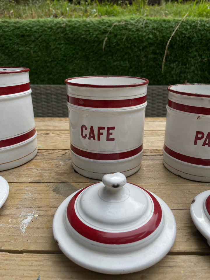 White enamel canisters with red stripes and the word 'CAFE' printed on them, arranged on a wooden table outdoors with a hedge in the background.