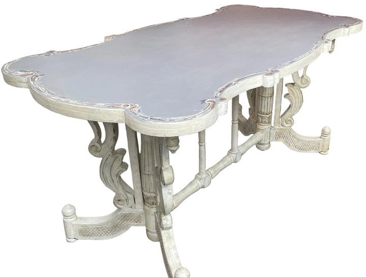 A distressed white carved dining table with a gray top, photographed on a tiled patio in an outdoor backyard setting next to a wicker sofa.
