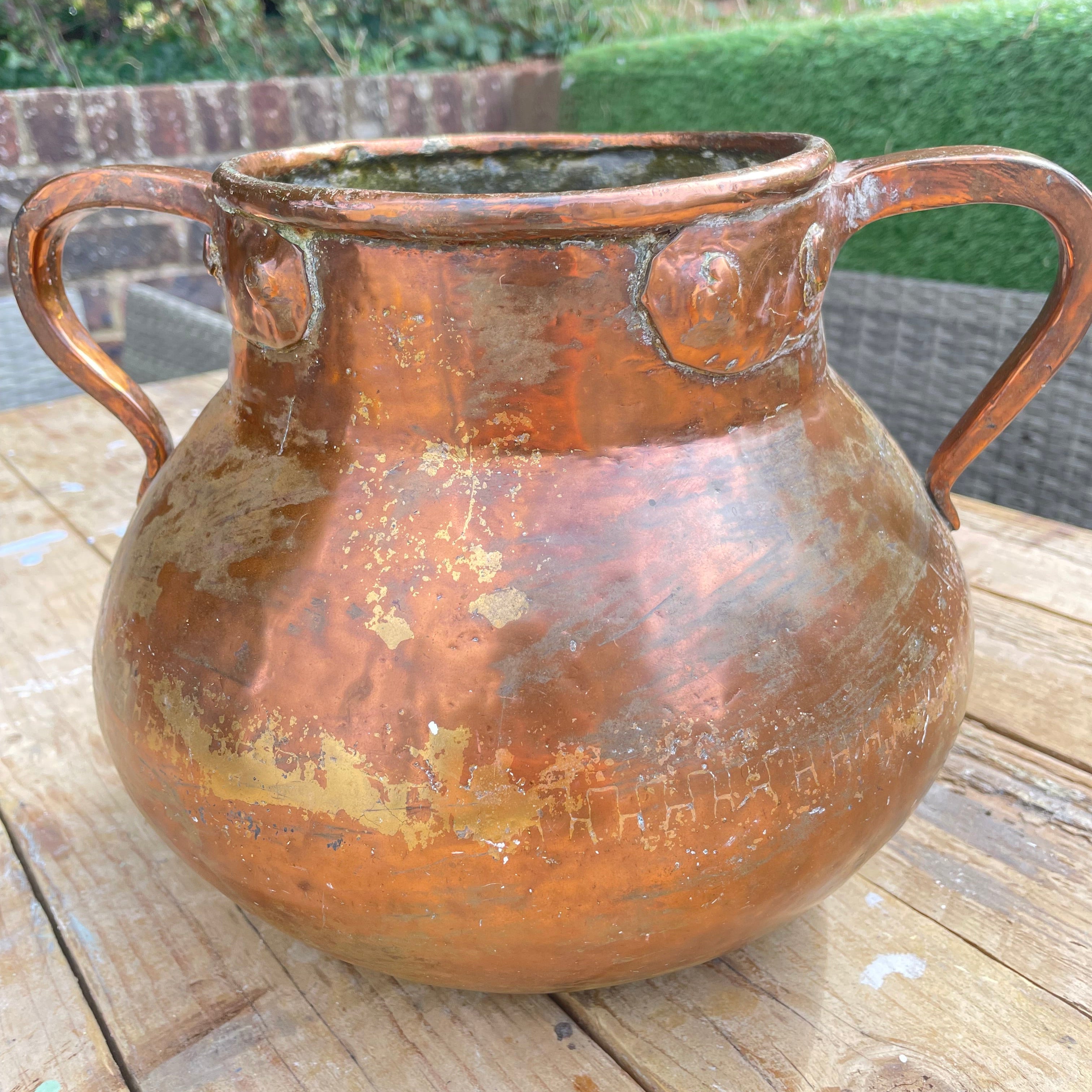 rustic copper-colored metal planter pot with two side handles, sitting on a wooden deck table with a garden background.