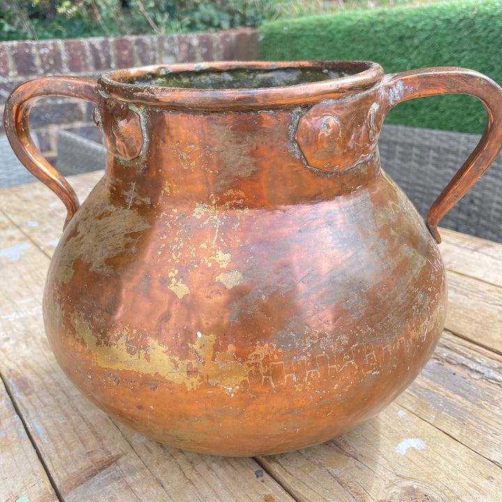 rustic copper-colored metal planter pot with two side handles, sitting on a wooden deck table with a garden background.