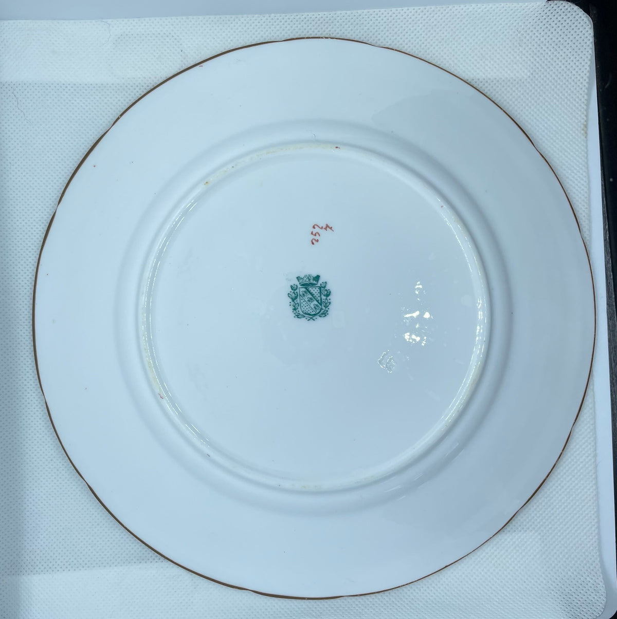 white porcelain plate with a thin gold rim, photographed from above inside a white protective box. The center of the plate features a small green floral mark and a small red handwritten number.
