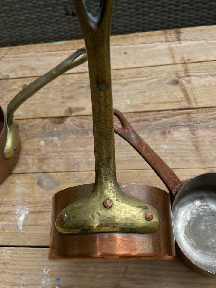 A vintage brass and copper pot holder stand holding a copper ladle, set on a rustic wooden table with other cookware partially visible.