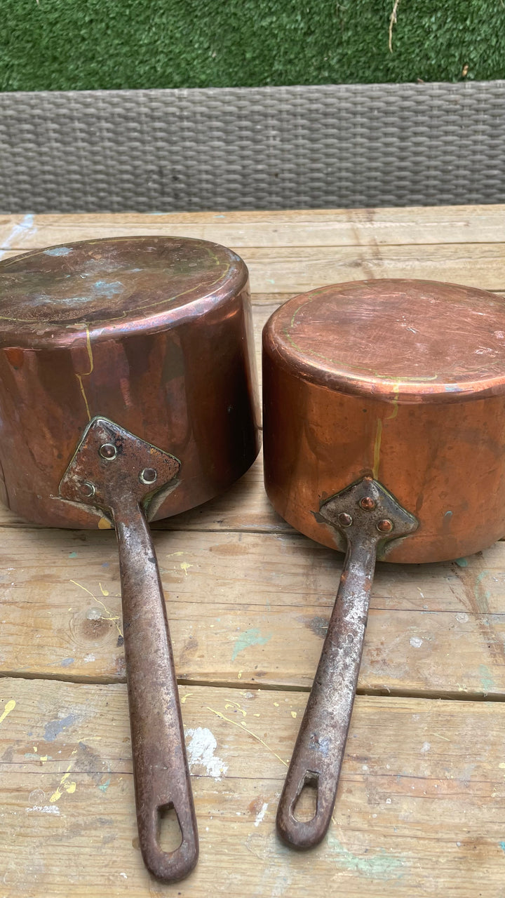 


Two vintage copper saucepans with long metal handles, resting on a wooden surface outdoors.