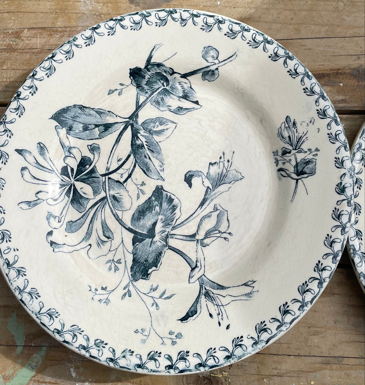 A round ceramic plate with a blue floral and leaf pattern on an off-white background, displayed on a rustic wooden surface.
