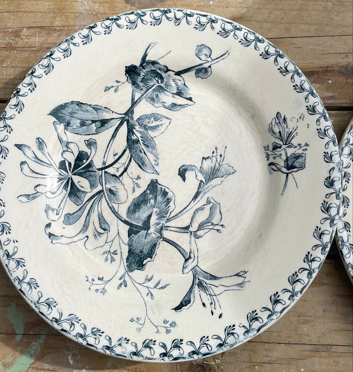 A round ceramic plate with a blue floral and leaf pattern on an off-white background, displayed on a rustic wooden surface.
