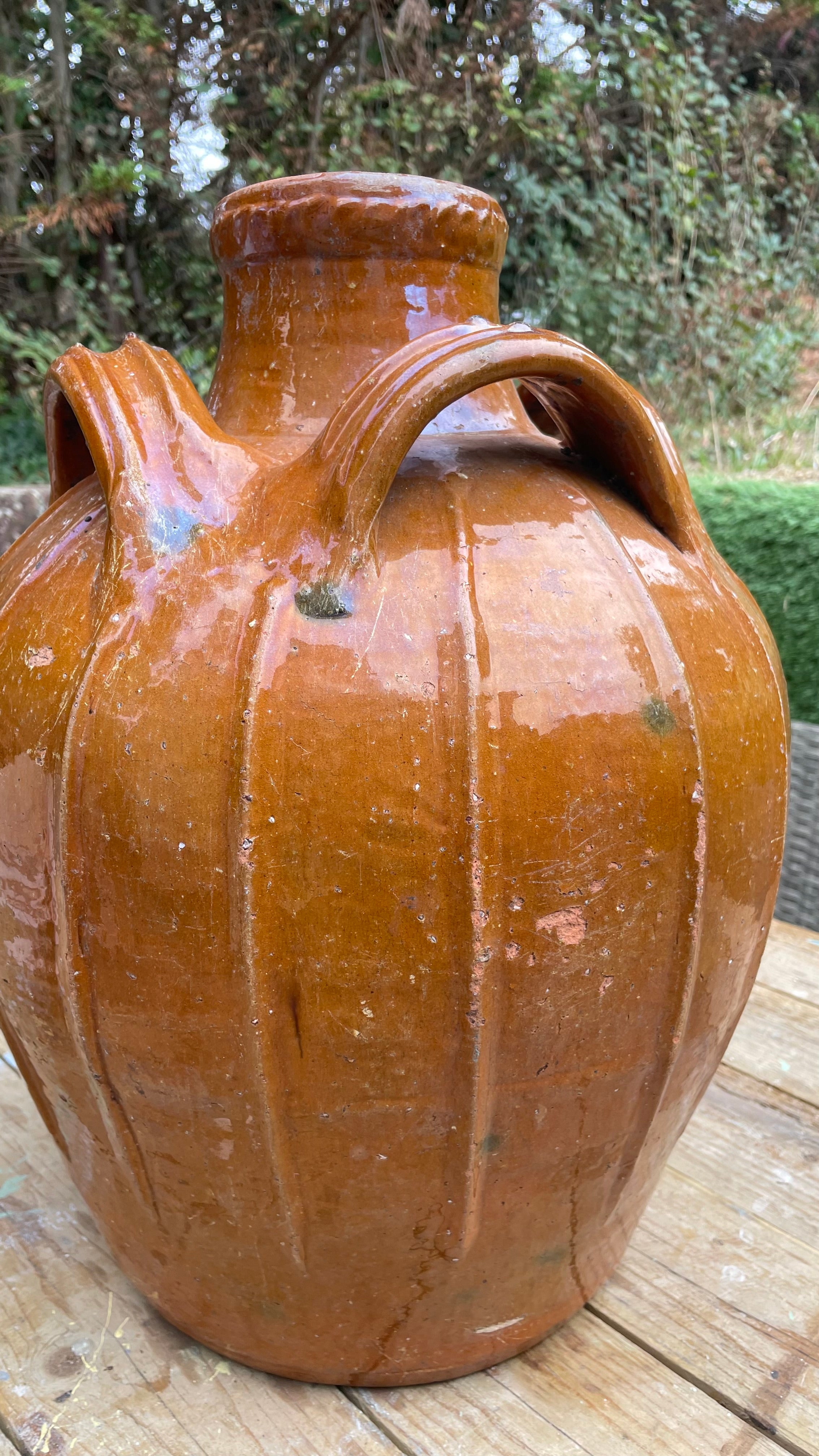 rustic brown glazed ceramic jug with a ribbed body, short neck, and two handles, shown on a wooden surface with an outdoor garden background.