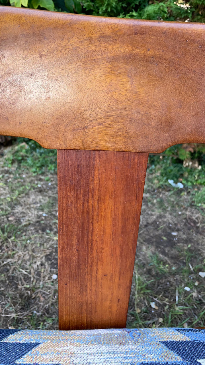close-up of a wooden chair back with a warm medium-brown finish, set outdoors with grass and foliage blurred in the background.