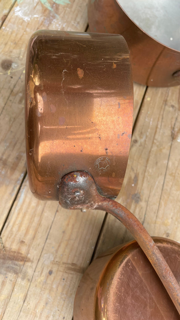 A close-up of a copper saucepan with a copper handle and lid, resting on a rustic wooden surface.