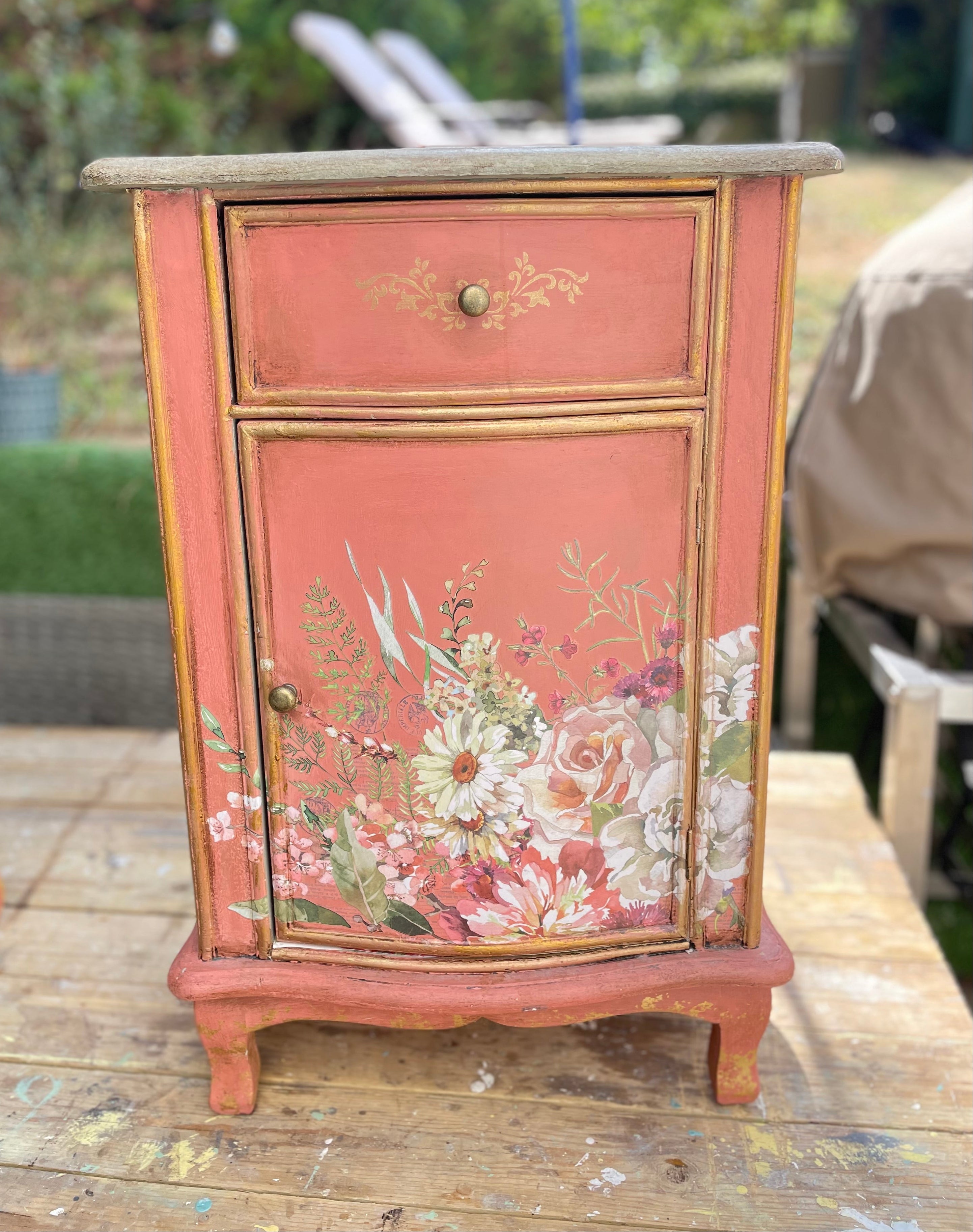small painted cabinet with a coral pink finish and gold trim, featuring floral artwork on the front door and a single drawer, photographed outdoors on a wooden surface with a garden background.