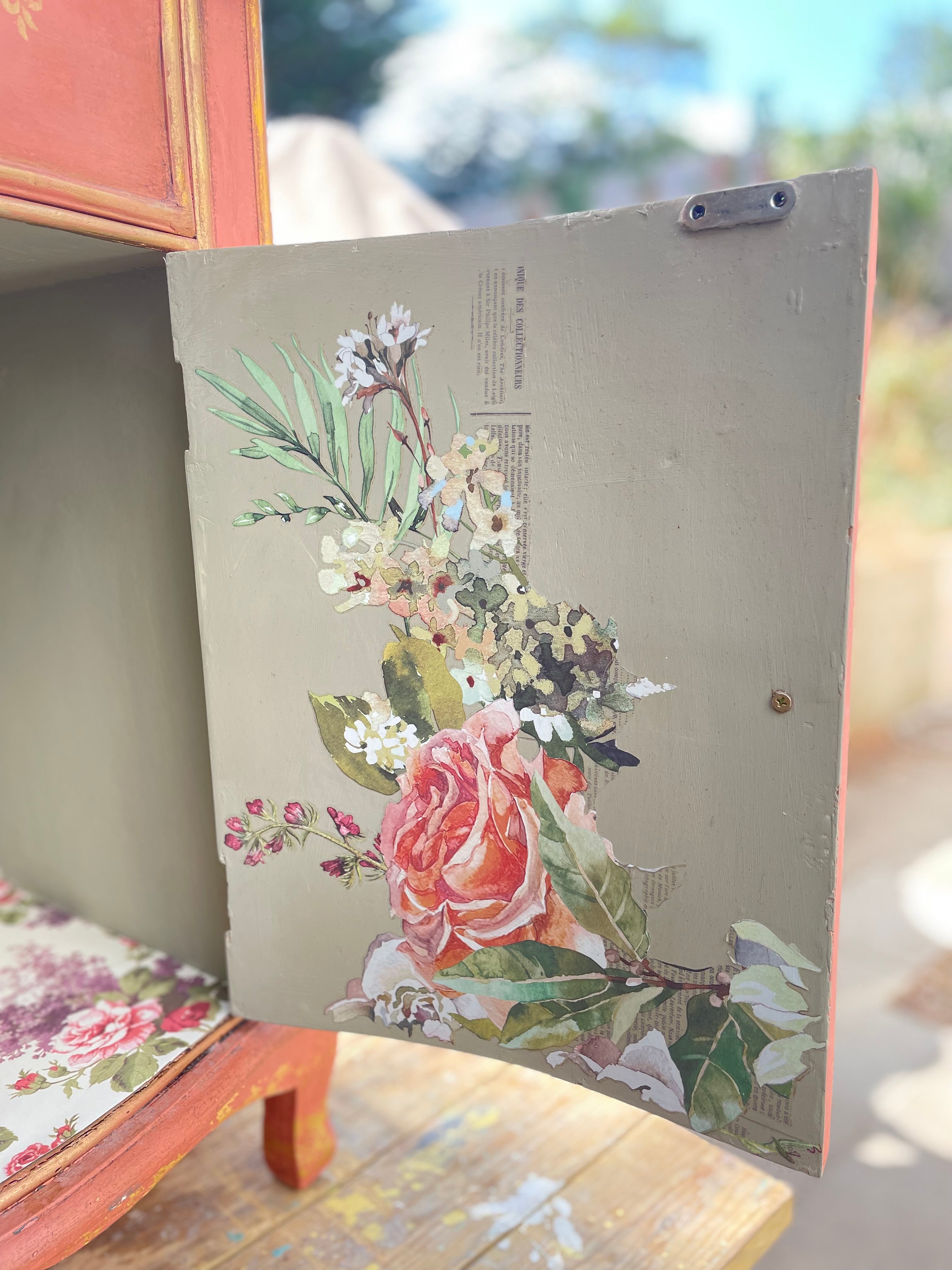vintage-style painted wooden board or panel with a floral print featuring a large pink rose and green leaves, leaning against furniture in an outdoor setting