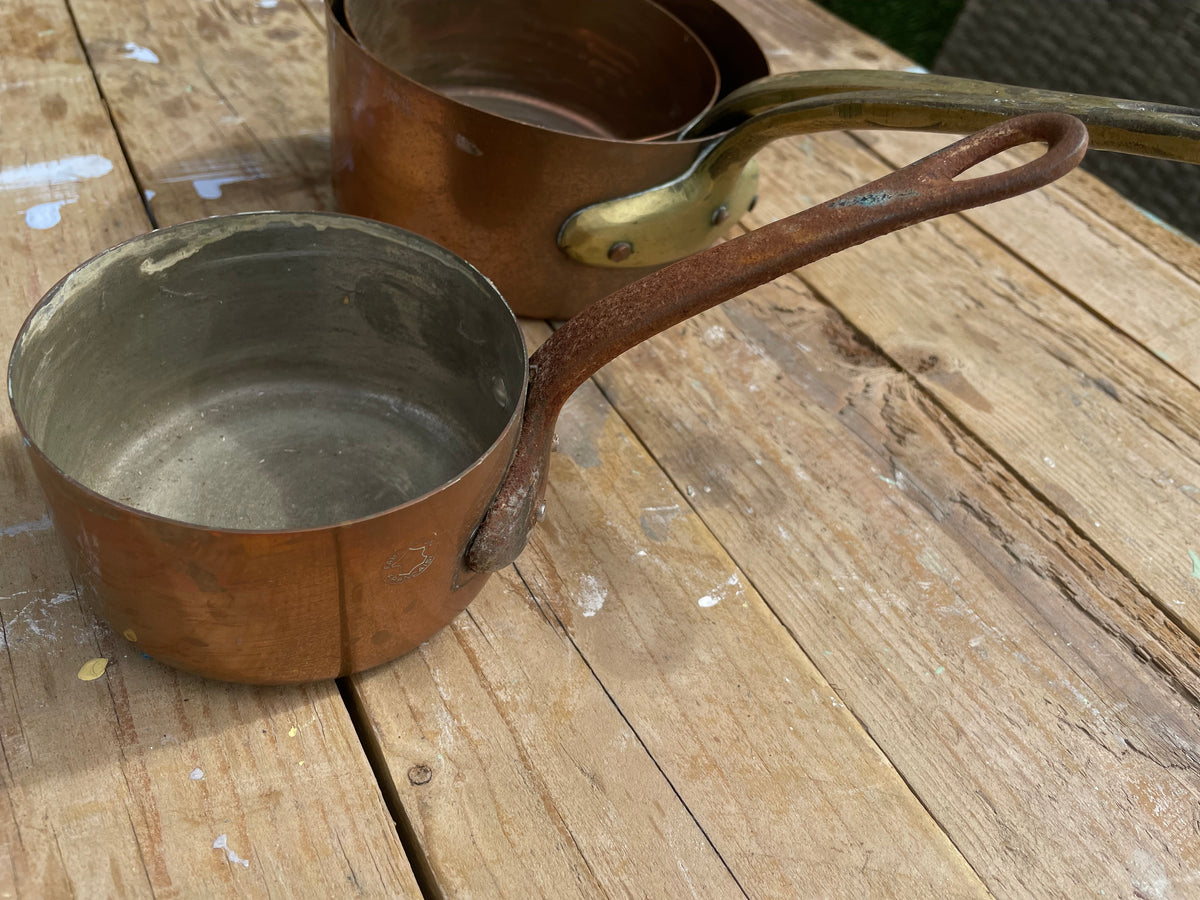 A set of vintage copper cookware pots with long handles and brass rivets, resting on a rustic wooden table.