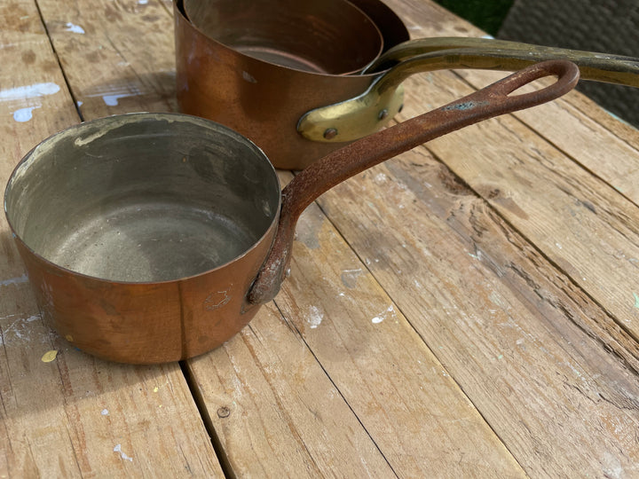 A set of vintage copper cookware pots with long handles and brass rivets, resting on a rustic wooden table.