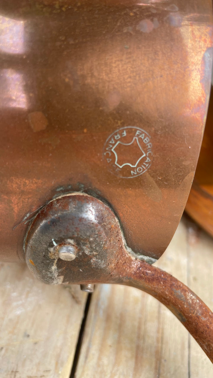 Close up of saucepan weathered copper surface, a rusted metal joint, and a stamped circular maker's mark on a light wood surface.