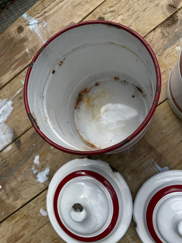 A close-up of a white enamel canister with a red rim, showing a stained interior, alongside matching white and red lids on a rustic wooden surface.