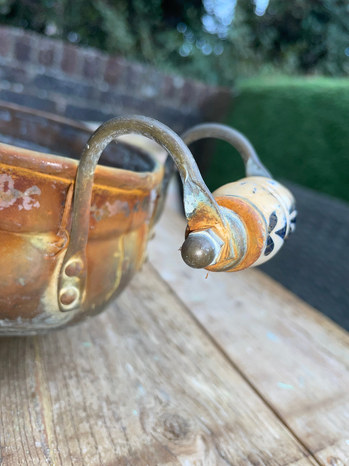 A rustic ceramic pitcher with a curved metal handle and a decorative cream, orange, and black patterned finial, shown on a wooden surface with an outdoor garden background.