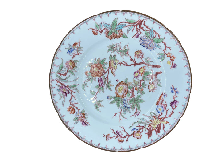 A round white porcelain plate with a colorful floral and fruit pattern, resting on a white textured surface inside an open box.