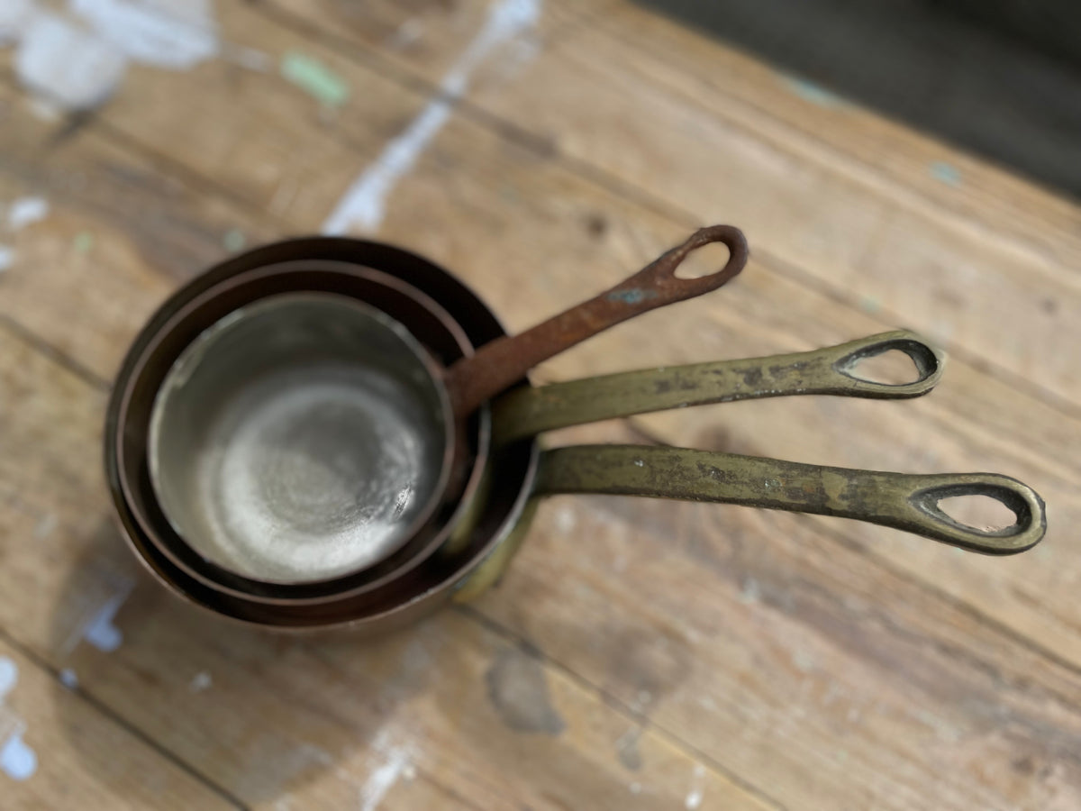 A set of nested vintage metal measuring cups with looped handles, nested inside a larger pot, resting on a rustic wooden surface.