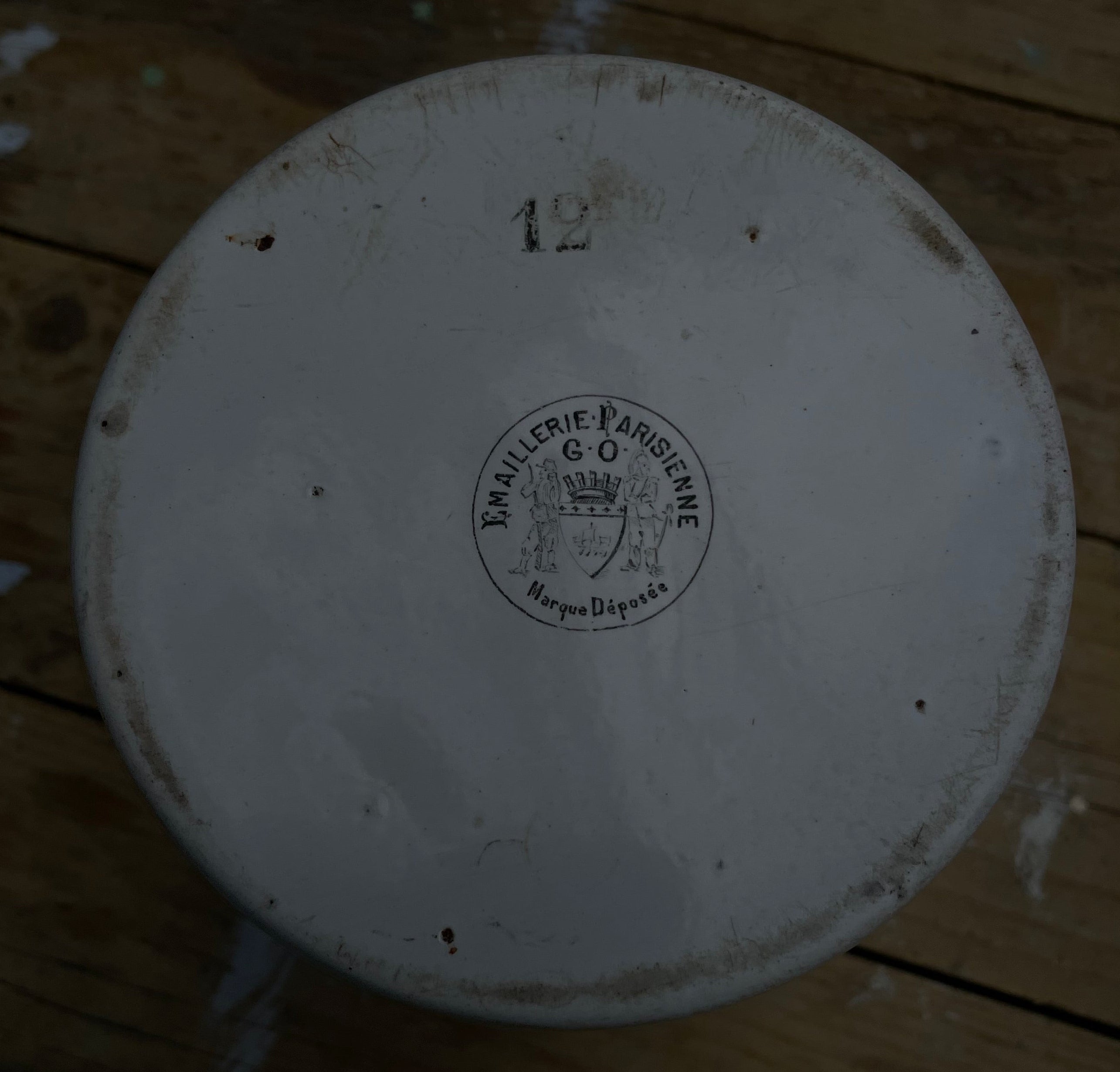 A top-down view of a round white ceramic plate or dish with a printed circular logo in the center and the number 12 near the top, placed on a weathered wooden floor.
