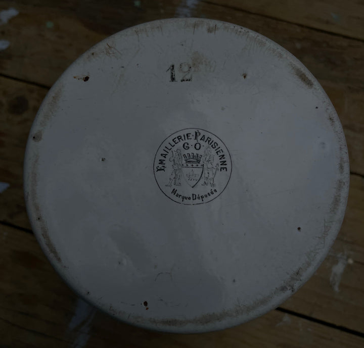 A top-down view of a round white ceramic plate or dish with a printed circular logo in the center and the number 12 near the top, placed on a weathered wooden floor.
