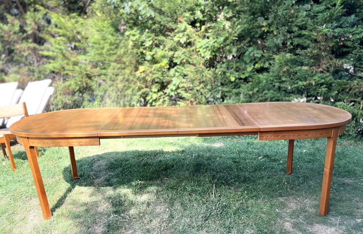 Wooden outdoor dining table with chairs on a grassy area with trees in the background By Troeds.
