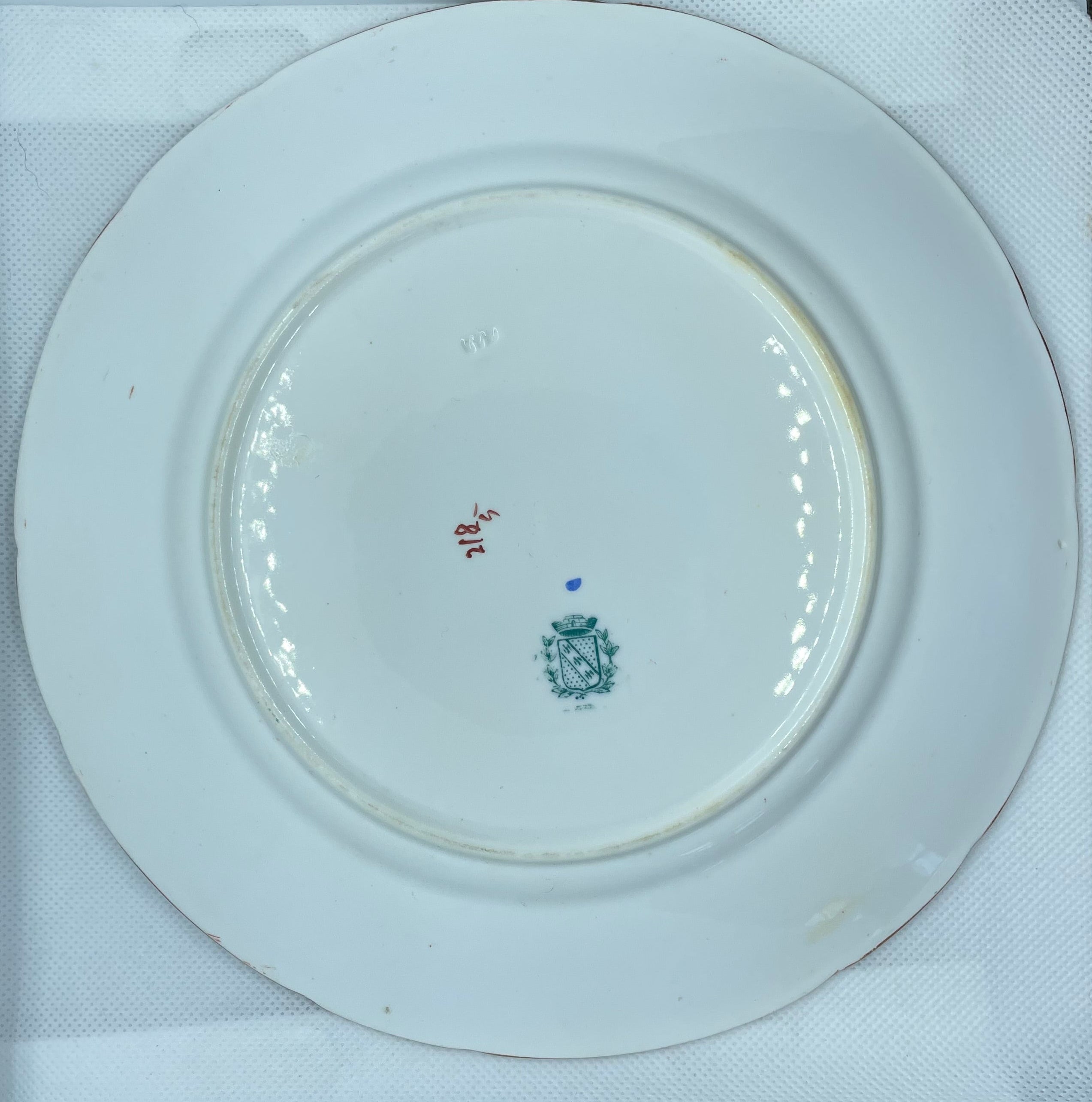 


A white porcelain plate with a small blue and red mark in the center, photographed from above inside a white box lined with white paper.