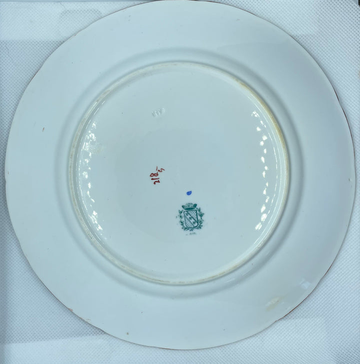 


A white porcelain plate with a small blue and red mark in the center, photographed from above inside a white box lined with white paper.