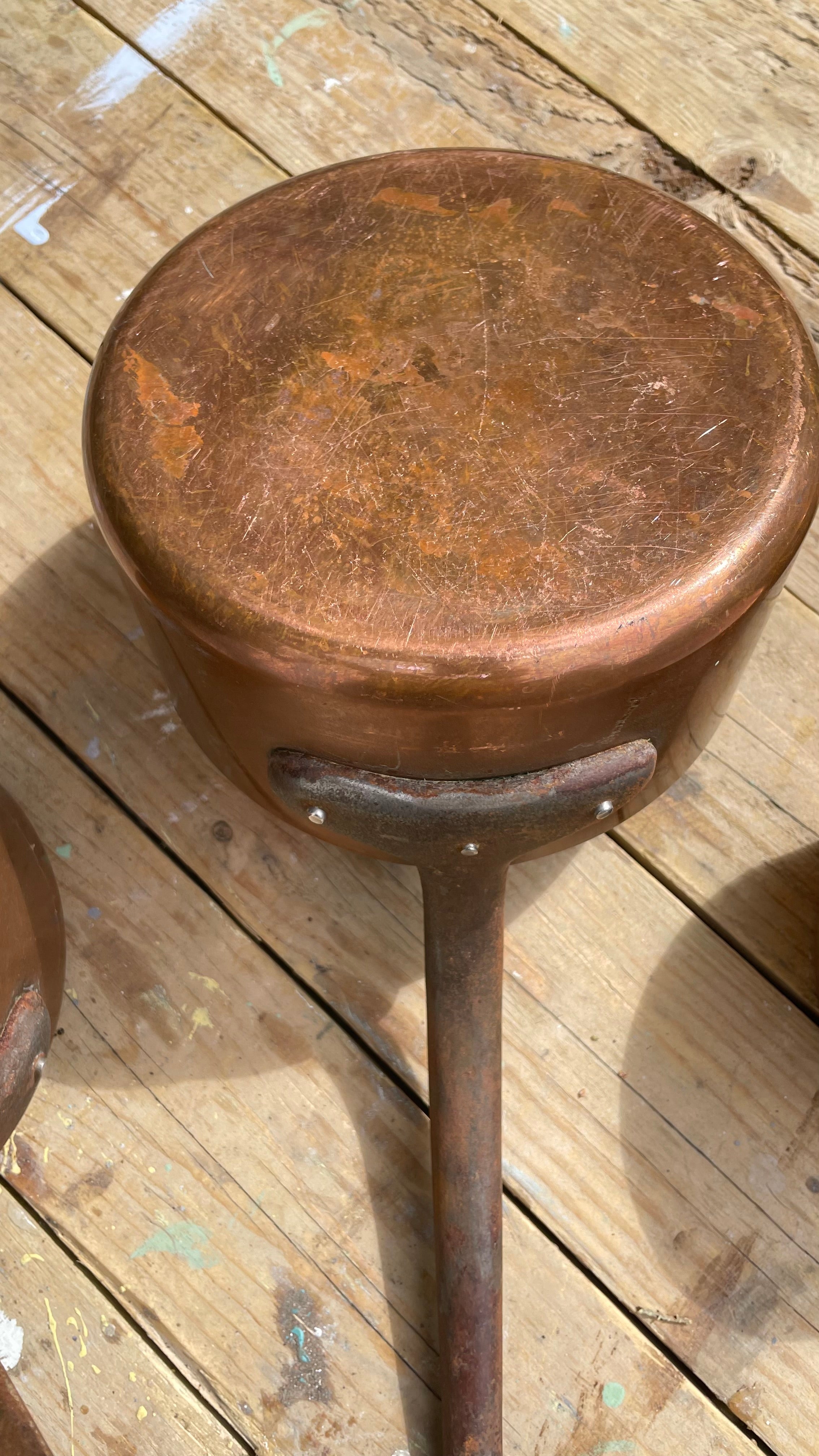 A vintage copper pot with a long handle and a round copper lid, resting on a wooden plank floor.