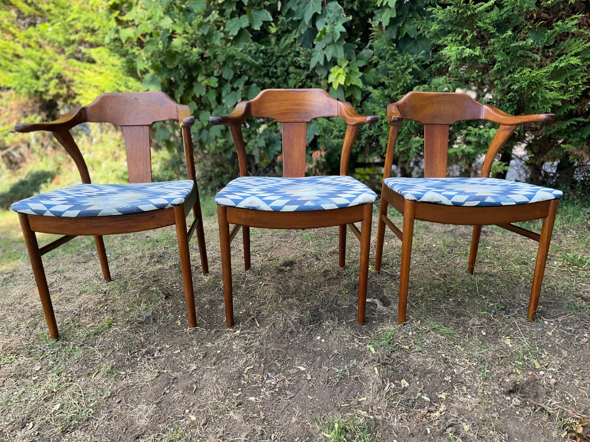 Three wooden armchairs with blue and white patterned seat cushions, arranged side-by-side on a dirt and grass lawn with green foliage in the background.